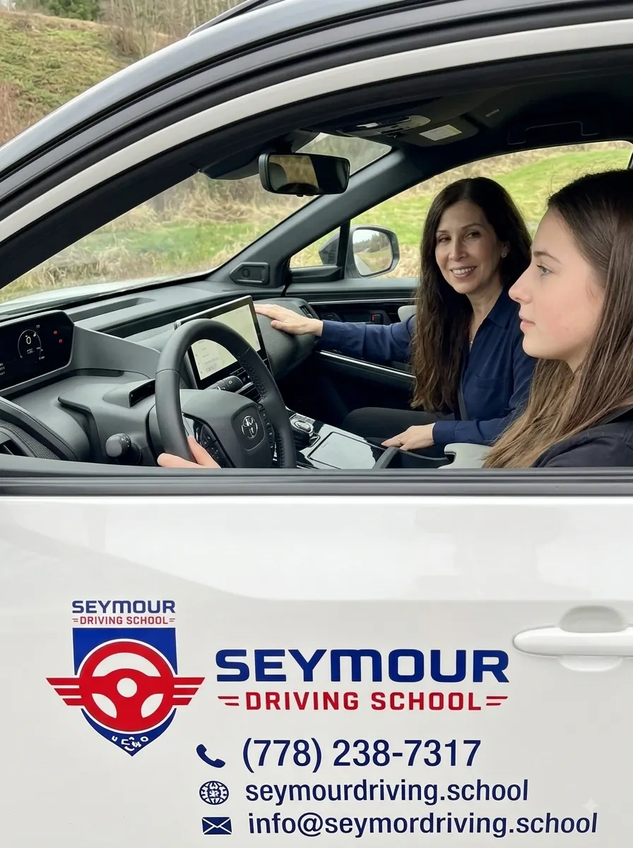 Inside a white Seymour Driving School car, a woman in a navy shirt points at the dashboard while a young driver holds the wheel.