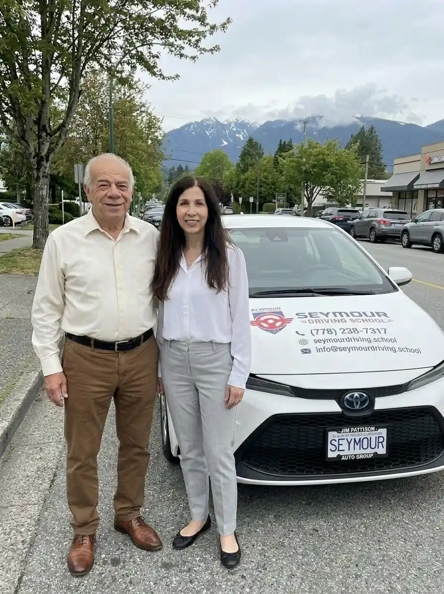 An older man in a white shirt and a woman in grey trousers stand in front of a white Seymour Driving School car.